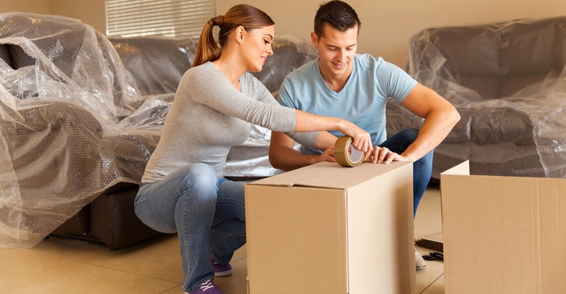 A couple packing items for storage at home, surrounded by furniture covered with clear dust covers.